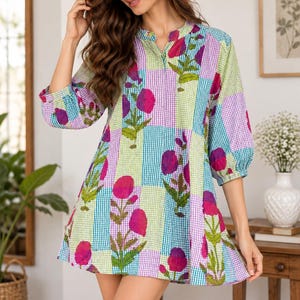 Op de afbeelding: Een tuniekjurk met korte mouwen en een levendig patchworkpatroon van blauwe, groene en witte vichy-ruitjes. Het kledingstuk is versierd met een herhalende bloemenprint van magentakleurige bloemen en groene stelen, afgewerkt met een opstaande kraag.