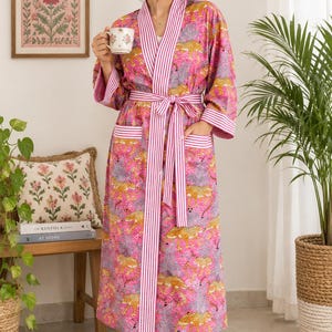 May include: A long-sleeved robe in a vibrant pink hue featuring a repeating leopard print pattern. The garment includes a white and pink vertical striped trim along the front opening, cuffs, and pockets, with a matching tie belt at the waist.