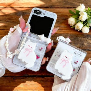 May include: Two clear foldable phone cases featuring white cat figures with pink bows, small cupcake and strawberry icons, and tiny heart accents. One case includes a white pearl beaded wrist strap with a small heart charm attached.