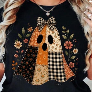 May include: A black t-shirt featuring a central graphic of a ghost made of various patchwork fabric patterns including orange, black, and white floral and plaid designs. The ghost is topped with a black and white plaid bow and flanked by floral stems.