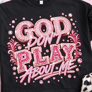 Op de afbeelding: Een zwart t-shirt met de tekst GOD DON'T PLAY ABOUT ME in een driedimensionaal, roze, glitterend lettertype in strass-stijl. Het ontwerp is omringd door roze bloemmotieven en verspreide roze ronde edelstenen op de voorkant van het shirt.