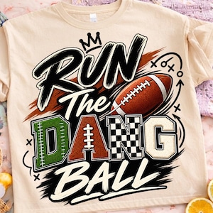 May include: A beige t-shirt featuring a graphic design with the text RUN The DANG BALL. The design includes a football, a crown icon, and play diagrams. The letters are styled with green, brown football texture, black and white checkers, and cream.