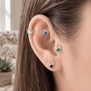 May include: Four silver-toned spider-shaped earrings are worn in a single ear. Each spider features a small, round gemstone in the center of its body. The gemstones are clear, blue, green, and black, respectively, set within the silver metal frames.