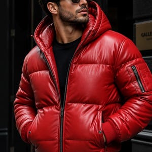 Könnte beinhalten: Eine leuchtend rote Daunenjacke mit Kapuze, Frontreißverschluss und einer Reißverschluss-Utility-Tasche am linken Ärmel. Die Jacke hat gerippte Bündchen und einen gerippten Saum, eine glatte Oberfläche und wird über einem schwarzen Shirt getragen.