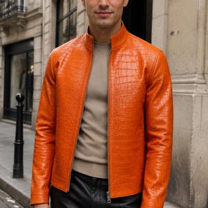 May include: A bright orange jacket with a textured crocodile-embossed pattern, featuring a stand-up collar, a full-length silver-tone metal zipper closure, and long sleeves. The jacket is worn over a beige crew-neck sweater and black trousers.