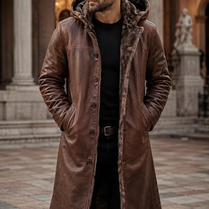 May include: A long brown leather coat featuring a hood lined with dark brown faux fur, a button-front closure, and long sleeves. The coat is worn over a black shirt and black pants, with the hands placed inside the side pockets of the garment.
