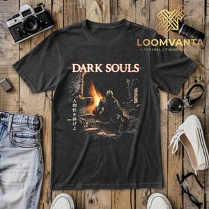 Może przedstawiać: Ciemnoszary t-shirt z krótkim rękawem z nadrukiem graficznym przedstawiającym rycerza siedzącego przy ognisku. Napis "DARK SOULS" jest nadrukowany na biało nad ilustracją, a japońskie znaki "人間性を捧げよ" znajdują się pionowo po lewej stronie.