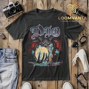 May include: A charcoal gray short-sleeve t-shirt featuring a graphic print of a horned demon figure holding a glowing moon. The text "DIO" is printed in a stylized font above the creature. The design includes red and blue color accents.