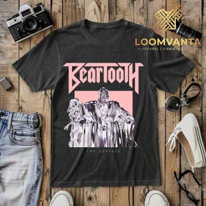 Puede incluir: Una camiseta gris carbón de manga corta con un estampado gráfico rosa claro. El diseño incluye la palabra Beartooth en una fuente estilizada sobre una figura de aspecto metálico sentada en un trono, con el texto The Surface debajo.