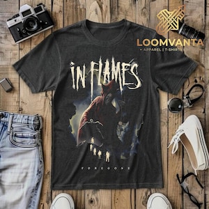 Puede incluir: Una camiseta gris carbón de manga corta con el texto IN FLAMES en una fuente blanca estilizada sobre un gráfico de una gran criatura roja y figuras más pequeñas. La palabra FOREGONE está impresa en letras blancas pequeñas en la parte inferior.