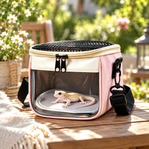 May include: A light pink and white pet carrier with a clear front window and a black mesh top. The carrier features black zippers and an adjustable black shoulder strap. Inside the carrier, a light-colored bearded dragon lizard rests on a gray mat.