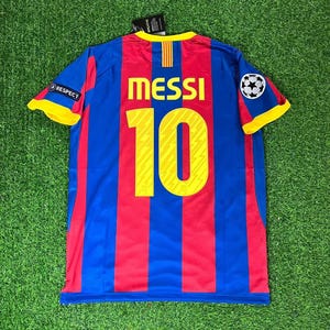 May include: A short-sleeved sports jersey featuring vertical red and blue stripes with yellow trim on the sleeves and collar. The back displays the name MESSI in yellow letters above the number 10, with a Respect patch and a football emblem.