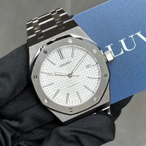 May include: A silver-toned Seiko automatic wristwatch featuring a textured white waffle-pattern dial, silver baton hour markers, and a date window at the three o'clock position. The watch has an octagonal bezel with exposed screws and a metal link band.