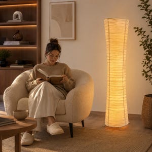 May include: A tall, cylindrical floor lamp with a textured, off-white paper shade that emits a warm, soft glow. The lamp features a slender, vertical structure with visible horizontal ribbing, standing on a simple metal base on the floor.