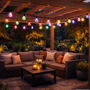 May include: A string of multi-colored LED globe lights hanging from a wooden pergola above an outdoor sectional sofa with gray cushions, decorative pillows, and a square coffee table featuring a lit candle and a small potted plant.