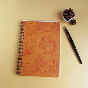 Peut inclure: Un carnet à spirales avec une couverture orange ornée d'un motif répétitif de hérissons, d'écureuils, de champignons et de baies en encre orange foncé. Un stylo-plume noir et deux petites pommes de pin brunes sont placés à proximité.