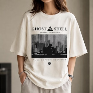 May include: An off-white short-sleeve t-shirt featuring a black graphic print of a city skyline and a silhouette figure. The text reads GHOST IN THE SHELL, THE NET IS VAST AND INFINITE, PUBLIC SAFETY SECTION 9, and includes a small square logo with text.