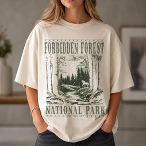 May include: A cream-colored t-shirt featuring a dark green graphic print of a forest landscape with a horse, a stream, and the text "FORBIDDEN FOREST NATIONAL PARK EST. 990 ADD" framed by rows of stars at the top and bottom of the design.
