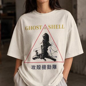 May include: An off-white short-sleeve t-shirt featuring a graphic print of a figure within a red triangle. The text reads GHOST IN THE SHELL in gold and Japanese characters in dark gray below the triangle. The design has a distressed aesthetic.