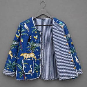 May include: A reversible quilted jacket featuring a blue exterior with a jungle print of leopards, toucans, and palm trees, and a white and blue vertical striped interior. The jacket has a round neckline, long sleeves, and a front button closure.