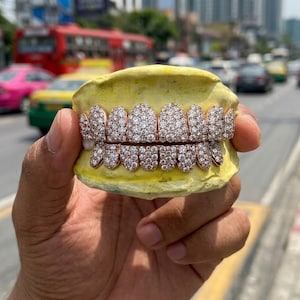 May include: A set of rose gold grillz featuring a dense pavé setting of clear, round-cut stones covering the entire surface of the upper and lower teeth. The dental jewelry is mounted on a yellow mold, showcasing a bright, reflective finish.