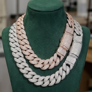 Może przedstawiać: Dwa grube łańcuchy o splocie typu cuban link, jeden w kolorze srebrnym, a drugi w kolorze różowego złota, oba w całości wysadzane małymi, przezroczystymi kamieniami. Każdy łańcuch posiada szerokie, prostokątne zapięcie typu box.