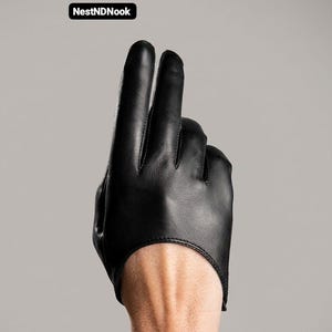 Puede incluir: Un guante de cuero negro que cubre solo el dedo índice y el medio, con un acabado liso y costuras visibles a lo largo de los dedos. El texto NestNDNook aparece en un pequeño recuadro negro en la esquina superior izquierda.
