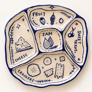 May include: A cream-colored ceramic serving platter with a wavy blue rim, divided into five sections. Each section features blue hand-drawn illustrations and labels: FRUIT, JAM, SWEET TREAT, CHEESE, and CRACKERS, decorated with small stars and vines.
