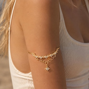 May include: A gold-toned arm cuff featuring a curved band set with small clear stones. A delicate chain drapes below the band, connecting to a central star-shaped charm that is also accented with a single clear stone in the center.