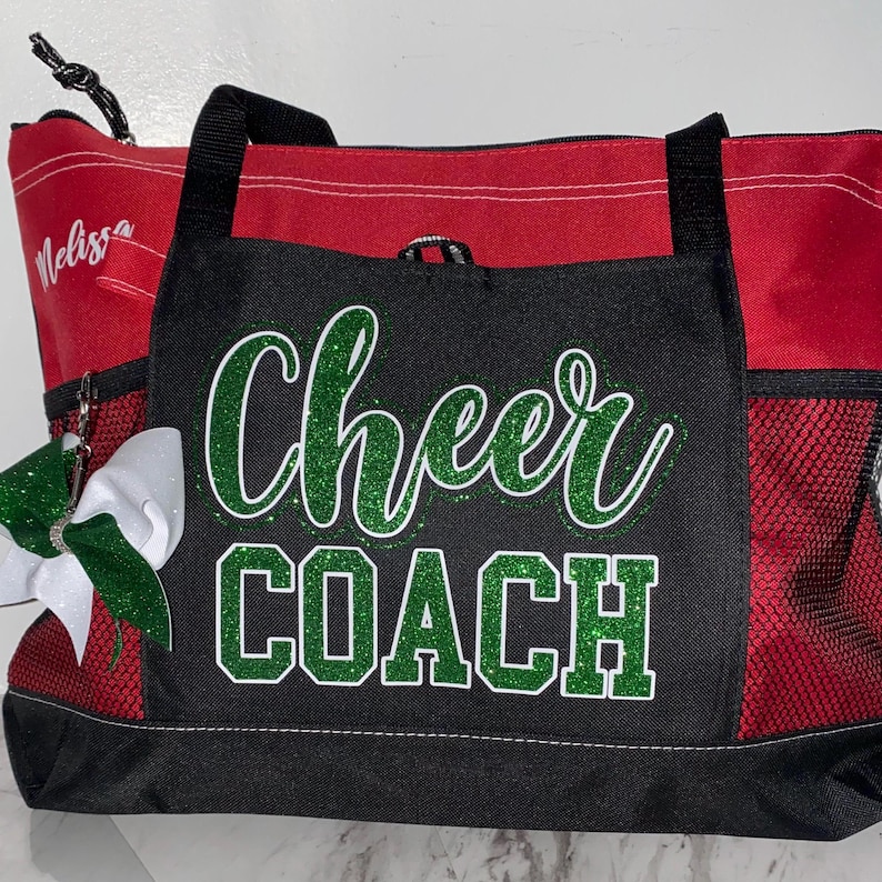 May include: Red and black tote bag with the words "Cheer COACH" in green glitter. The bag has black handles and a side pocket. A white and green bow is attached to the side. The name "Melissa" is written on the side.