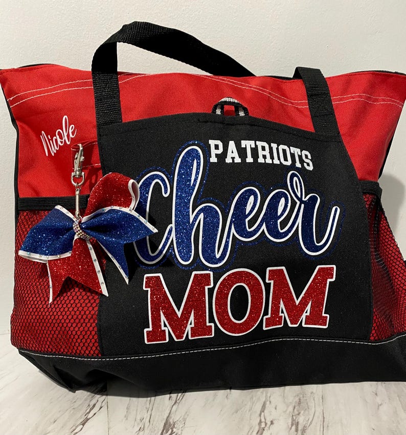 May include: Red and black tote bag with "Patriots Cheer Mom" text in blue and red glitter. Features a red and blue bow with a silver clip. Includes a side pocket and a black handle.