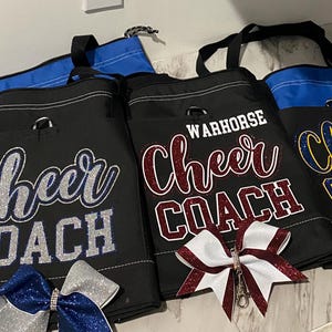 May include: Black tote bags with "Cheer COACH" and "WARHORSE Cheer COACH" text in glittery lettering. The bags have blue accents and are paired with cheer bows in silver, blue, and burgundy. The bags are designed for cheerleading coaches.