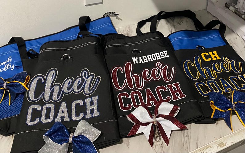 May include: Black tote bags with "Cheer COACH" and "WARHORSE Cheer COACH" text in glittery lettering. The bags have blue accents and are paired with cheer bows in silver, blue, and burgundy. The bags are designed for cheerleading coaches.