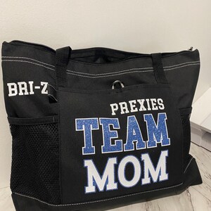 May include: Black tote bag with the words "PREXIES TEAM MOM" in blue and white lettering. The bag has a zippered top, side pockets, and a handle. The name "BRI-Z" is printed in white on the side of the bag.