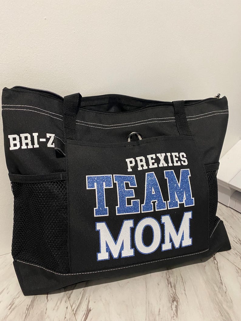May include: Black tote bag with the words "PREXIES TEAM MOM" in blue and white lettering. The bag has a zippered top, side pockets, and a handle. The name "BRI-Z" is printed in white on the side of the bag.