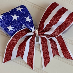 May include: A red, white, and blue glitter cheer bow with a star-spangled design. The bow is tied with a white ribbon and has a rhinestone center.
