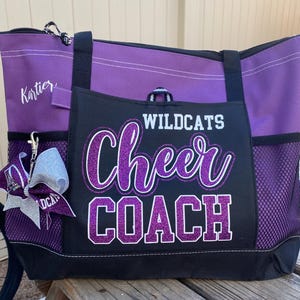 May include: Purple and black tote bag with the words "Wildcats Cheer Coach" in glittery purple lettering. The bag has a zippered top, side pockets, and a silver bow keychain.