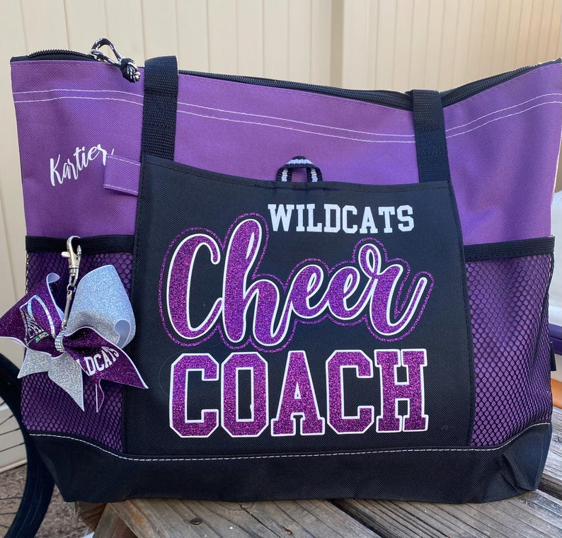 May include: Purple and black tote bag with the words "Wildcats Cheer Coach" in glittery purple lettering. The bag has a zippered top, side pockets, and a silver bow keychain.