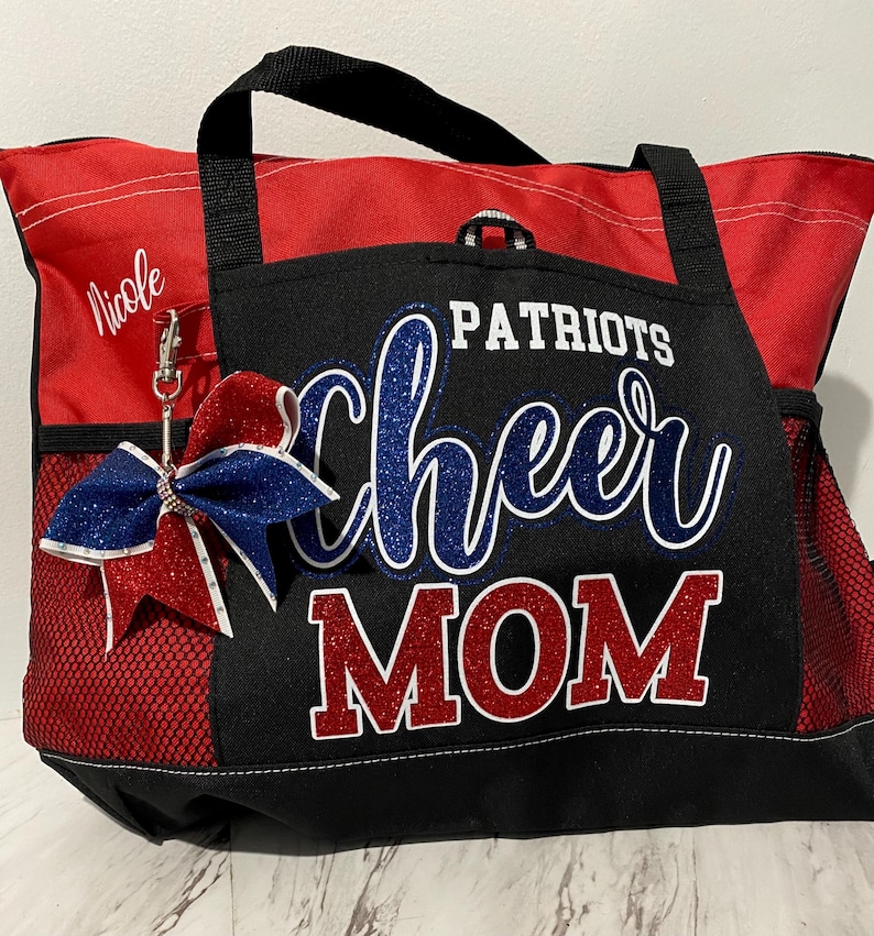 May include: Red and black tote bag with "Patriots Cheer Mom" text in blue and red glitter. Features a red and blue bow with a silver clip. Includes a side pocket and a black handle.