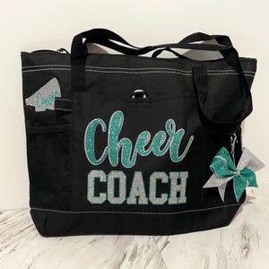 May include: Black tote bag with the words "Cheer COACH" in teal and silver glitter. The bag has a zipper closure, multiple pockets, and a teal and silver bow charm. A megaphone graphic is on the side.