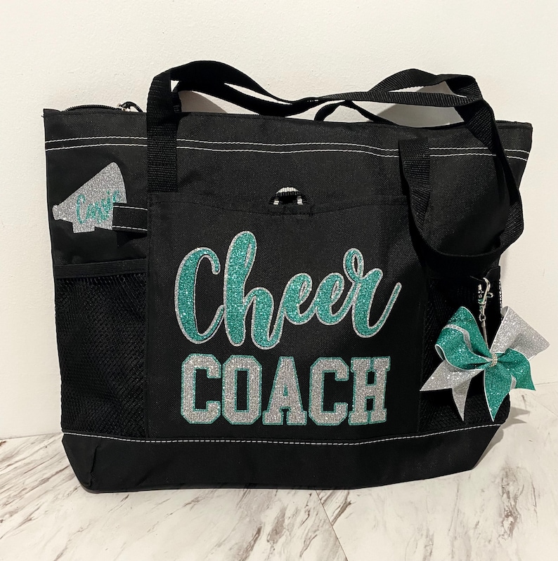 May include: Black tote bag with the words "Cheer COACH" in teal and silver glitter. The bag has a zipper closure, multiple pockets, and a teal and silver bow charm. A megaphone graphic is on the side.