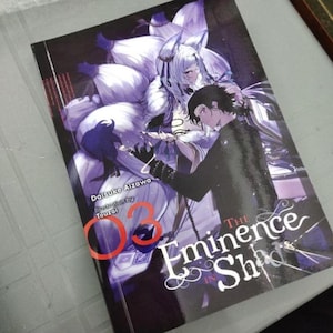 May include: A manga book titled The Eminence in Shadow volume 03 by Daisuke Aizawa with illustrations by Touzai. The cover features a character with long white hair and multiple tails behind a dark-haired character in a black outfit.
