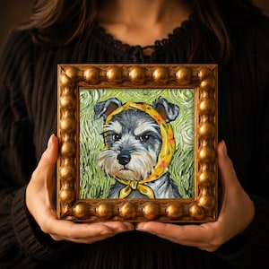 May include: An oil painting of a grey and white schnauzer dog wearing a yellow headscarf with small red floral patterns. The painting is set within a square, metallic gold frame featuring a decorative, rounded beaded border design.