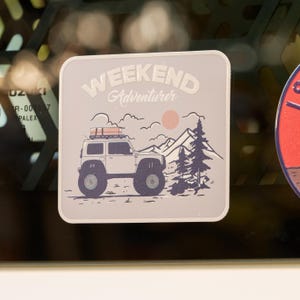 Op de afbeelding: Een vierkante sticker met afgeronde hoeken, een lichtgrijze achtergrond en een donkerblauwe illustratie van een terreinwagen met imperiaal, dennenbomen, bergen en een lichtroze zon. De tekst luidt WEEKEND Adventurer.