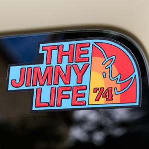 May include: A rectangular sticker with a light blue background and a black border. The text THE JIMNY LIFE is printed in bold red letters. To the right, a semi-circular graphic features a sunset design in yellow, orange, and red with the number 74.