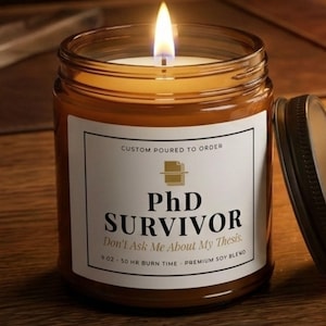 May include: A lit candle in an amber glass jar with a black metal lid. The white label features the text: CUSTOM POURED TO ORDER, PhD SURVIVOR, Don't Ask Me About My Thesis, 9 OZ - 50 HR BURN TIME - PREMIUM SOY BLEND, and a book icon.