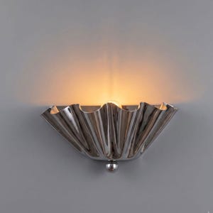 Peut inclure: Une applique murale en chrome poli dotée d'un abat-jour en forme d'éventail plissé semi-circulaire avec une surface métallique réfléchissante. Une ampoule est placée au centre, diffusant une lueur chaude sur le mur au-dessus du luminaire.