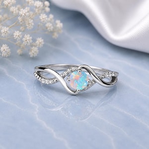 Peut inclure: Une bague argentée ornée d'une pierre centrale ronde en opale irisée. L'anneau présente un design torsadé et croisé, agrémenté de petites pierres claires et scintillantes sur les côtés ainsi que de perles métalliques près du sertissage.