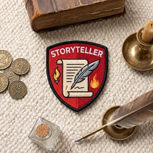 May include: A shield-shaped embroidered patch with a red background and black border. The text STORYTELLER is in white at the top. The center features a beige scroll, a grey quill pen, and two small orange and yellow flames on either side.