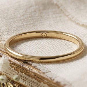 Op de afbeelding: Een dunne, gepolijste gouden ring met de gegraveerde letters KA aan de binnenzijde. De ring heeft een glad, afgerond profiel en een reflecterende metalen afwerking, wat een eenvoudig en strak ontwerp toont met duidelijke, leesbare letters.